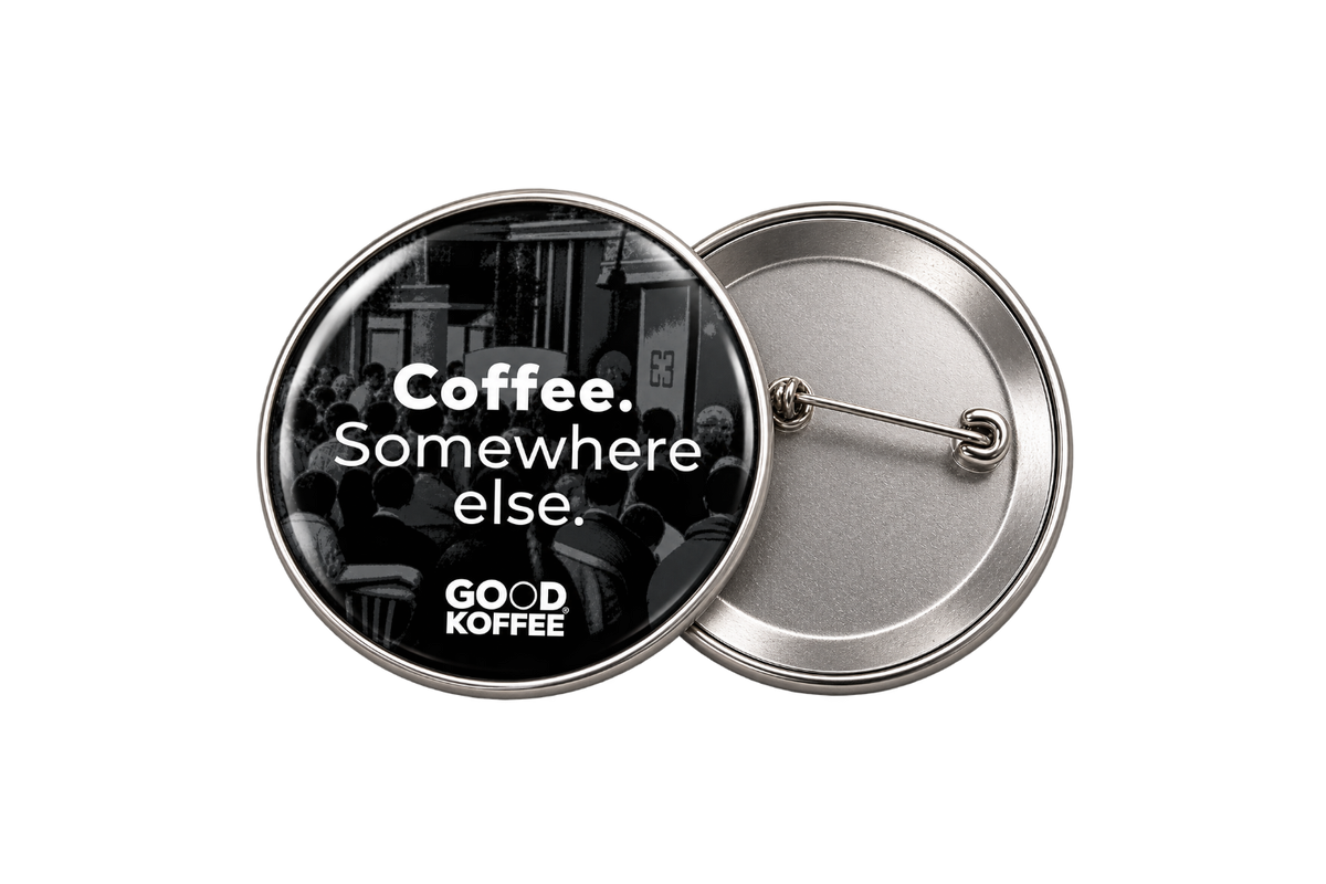 Two round metal pinback buttons shown on a white background. The front button has a black-and-white photo of a crowd with the text 'Coffee. Somewhere else'.