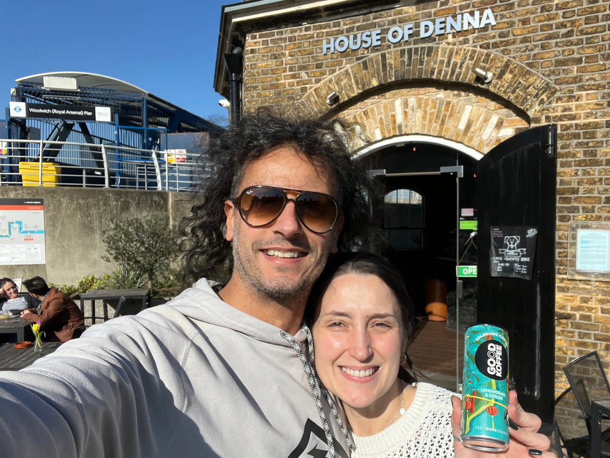 A man and a woman taking a selfie outdoors in front of a brick building with 'HOUSE OF DENNA' visible on the facade. The man is wearing sunglasses and a gray hoodie, and the woman is wearing a white top.