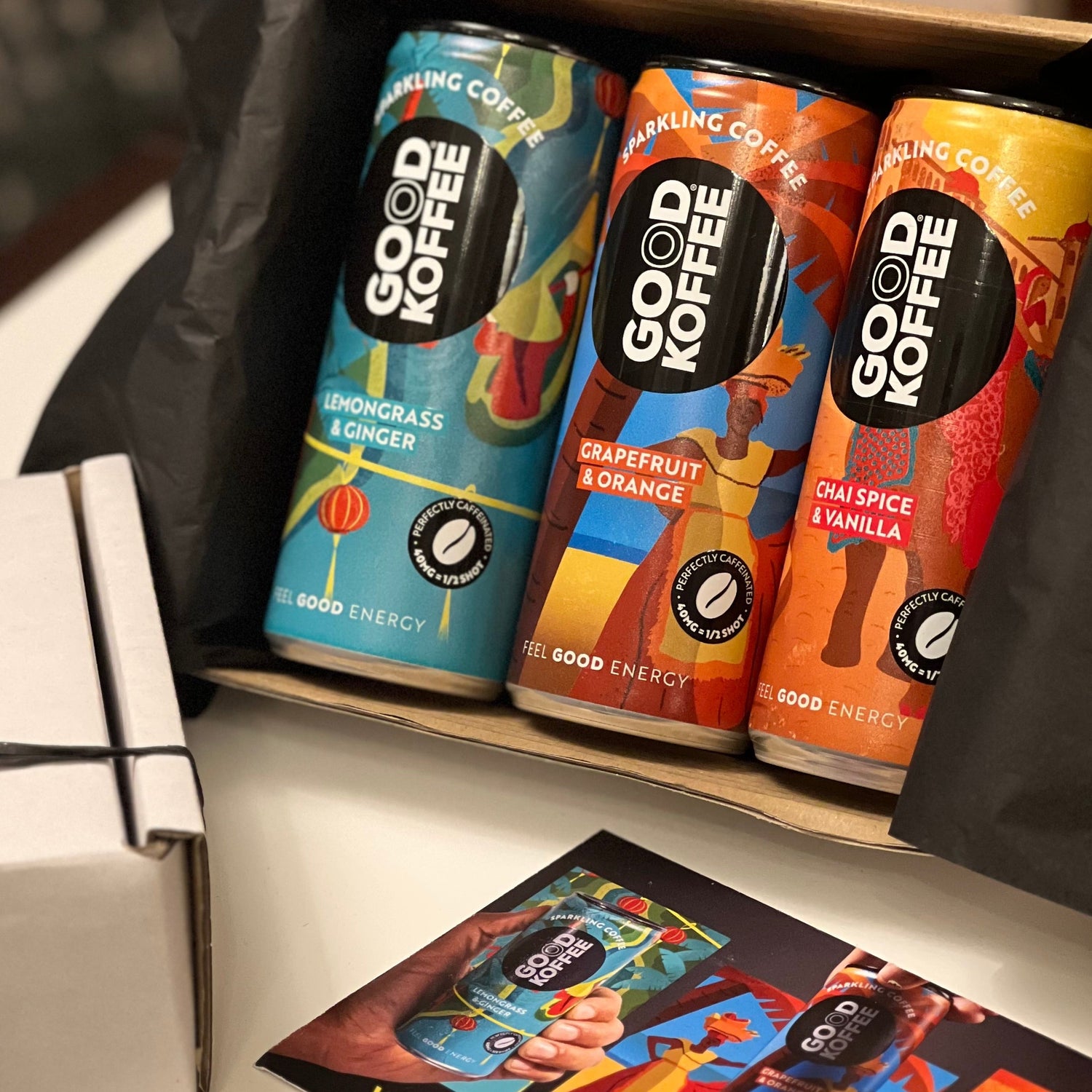 3 Cans in a white display box with a GOOD KOFFEE postcard sitting in front