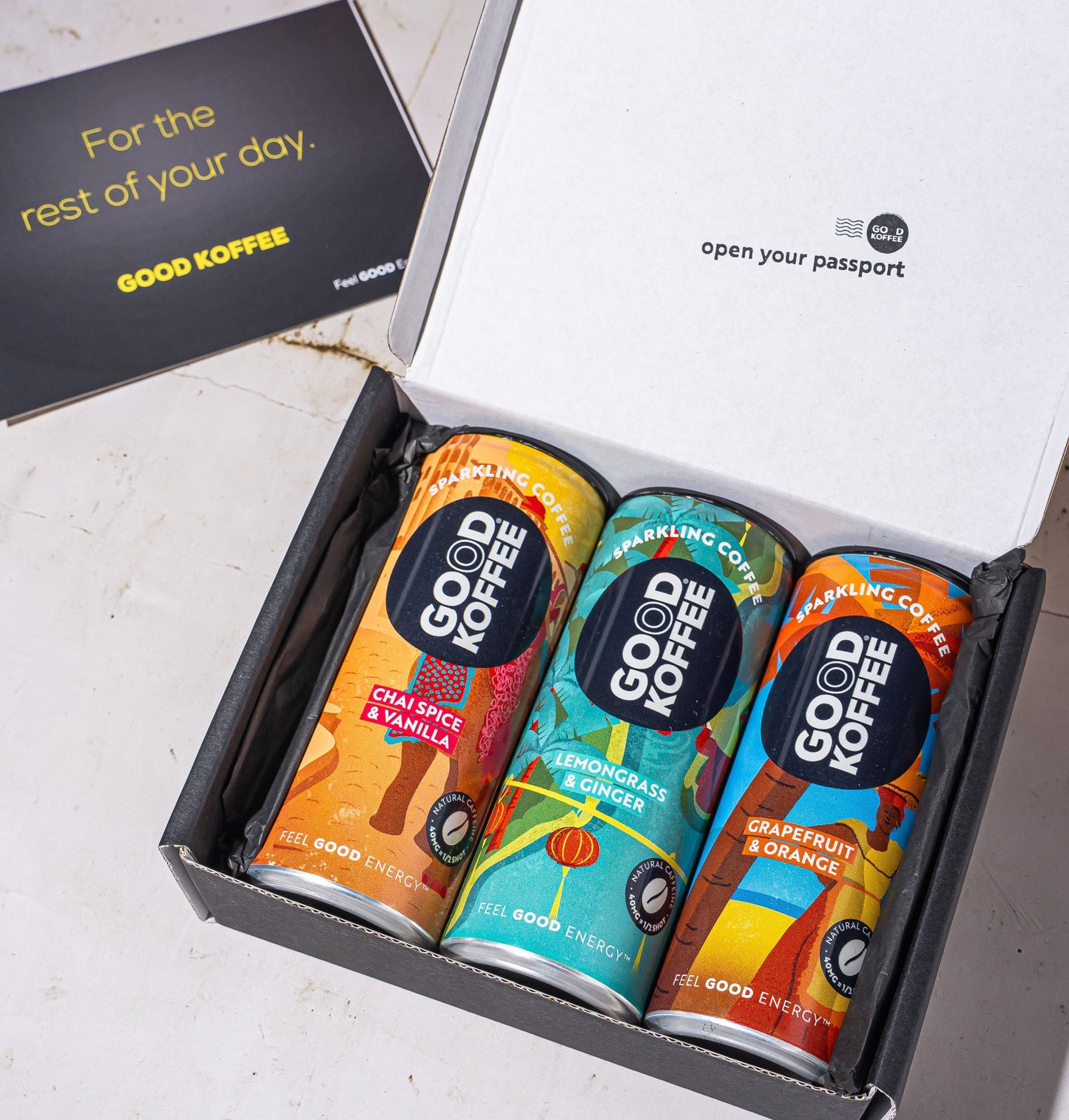 An open black gift box containing three colorful Good Coffee cans, with a black card beside it on a light stone surface. The inside lid reads 'open your passport' and the card says 'For the rest of your day.