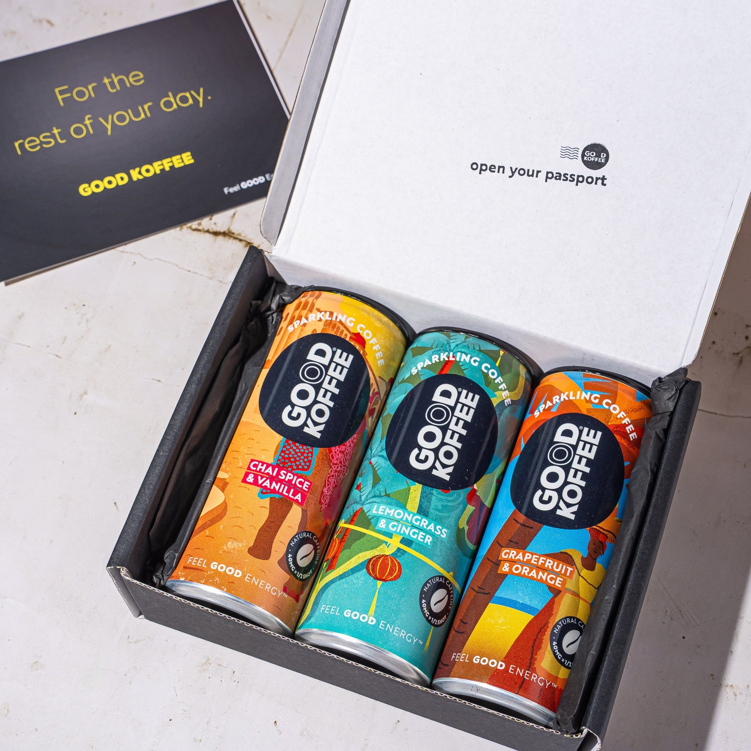 An open black gift box containing three colorful Good Coffee cans, with a black card beside it on a light stone surface. The inside lid reads 'open your passport' and the card says 'For the rest of your day.