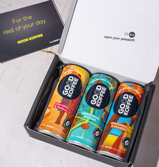 An open black gift box containing three colorful Good Coffee cans, with a black card beside it on a light stone surface. The inside lid reads 'open your passport' and the card says 'For the rest of your day.