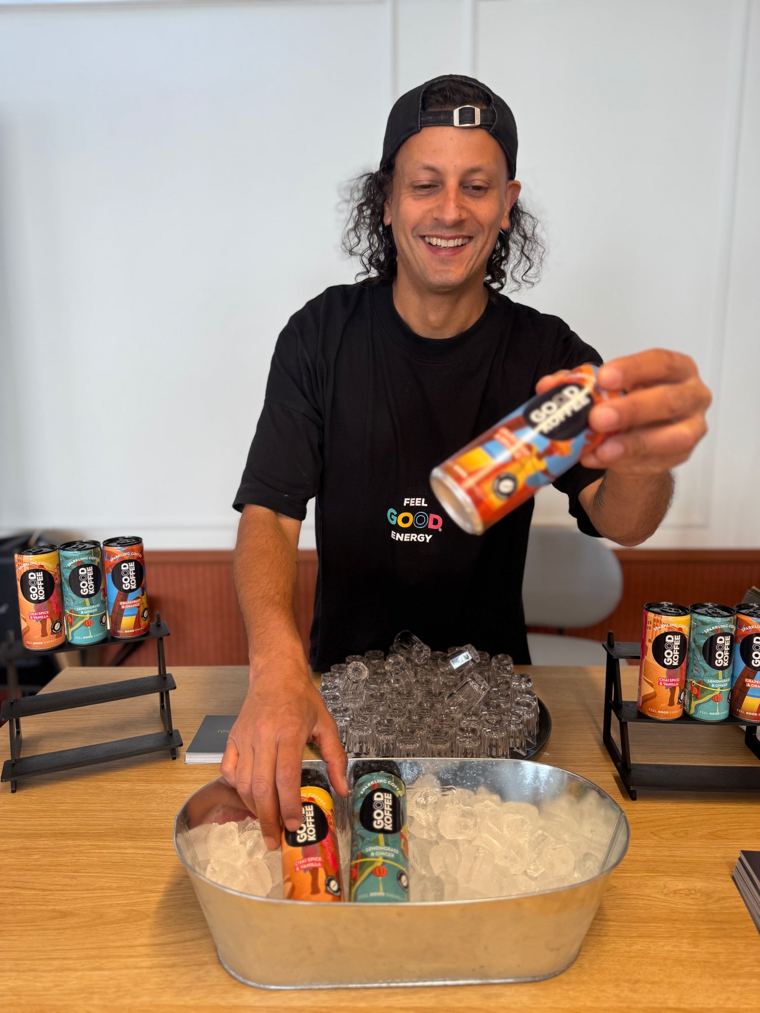 A smiling man wearing a black t-shirt and a black cap stands behind a table filled with ice, holding a colorful can of Sober Energy. Several cans of Sober Energy are displayed on stands on both sides, and two cans are placed in the metal tub of ice in front of him.