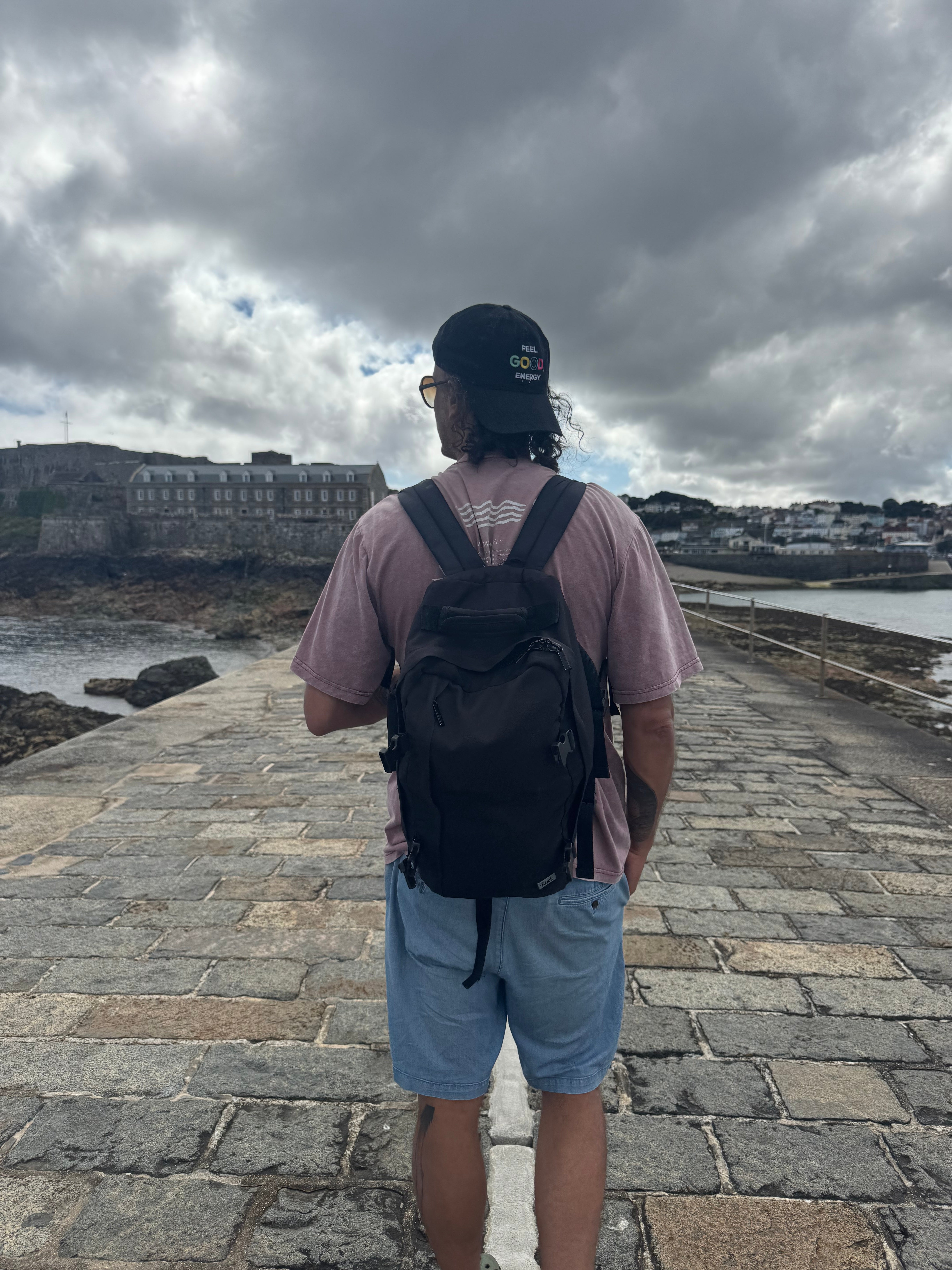 A man viewed from behind wearing a black backpack, a dusty pink t-shirt, and light blue shorts, standing on a stone pier with a coastal town and cloudy sky in the background.