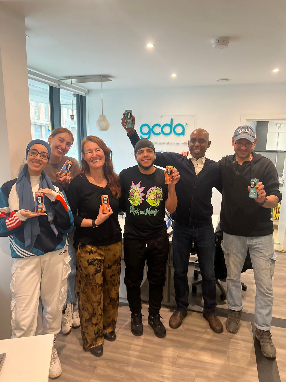 A group of seven people posing together in an office environment, each holding up a can of GOOD KOFFEE sparkling coffee. The background features a white wall with the logo 'gcda' in blue.
