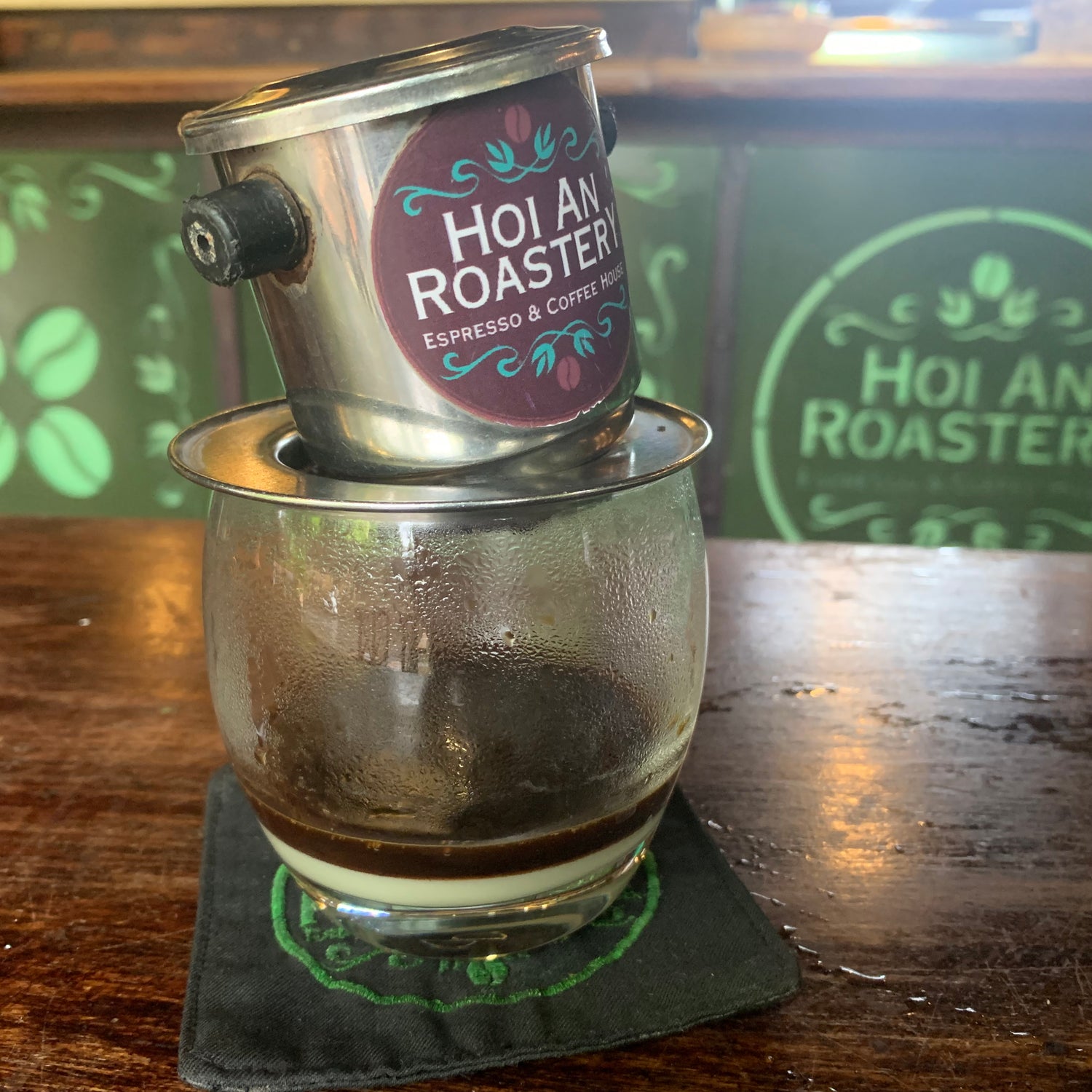 A Hoi An Roaster espresso maker sitting on a dark wooden bar counter, with brewed coffee dripping into a clear glass cup. The background features green wood paneling with the Hoi An Roaster logo.