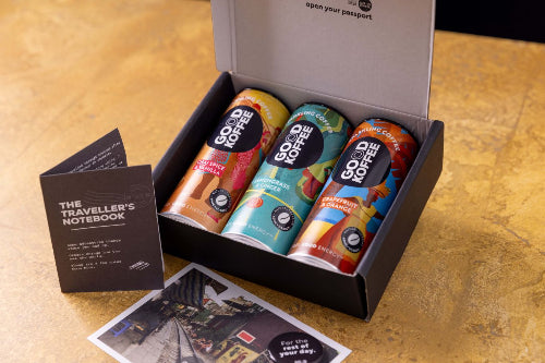 A gift set containing three GOOD KOFFEE cans in a black gift box, displayed on a textured gold tabletop next to a black notebook and a passport photo print.