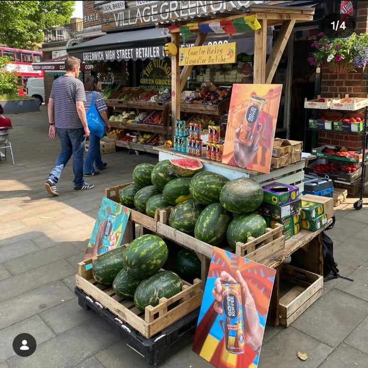 A social media post screenshot showing a street market scene with wooden crates filled with watermelons in the foreground, a vendor stand, and people walking in the background. The post header reads 'goodkoffeedrink and villagegreengrocers' and includes comments below.