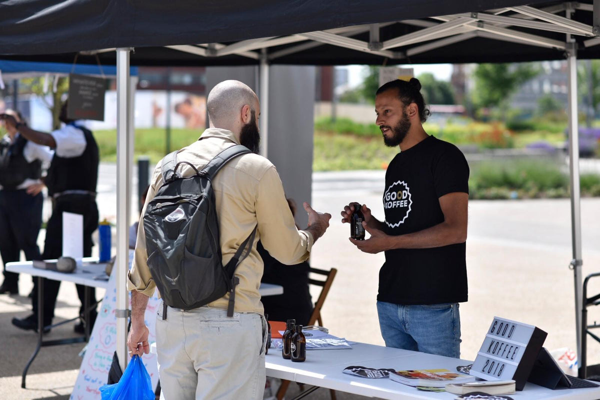 A man in a black t-shirt with a white circular logo reading 'GOOD KOFFEE' hands a bottle to a customer under a black canopy tent at an outdoor event.