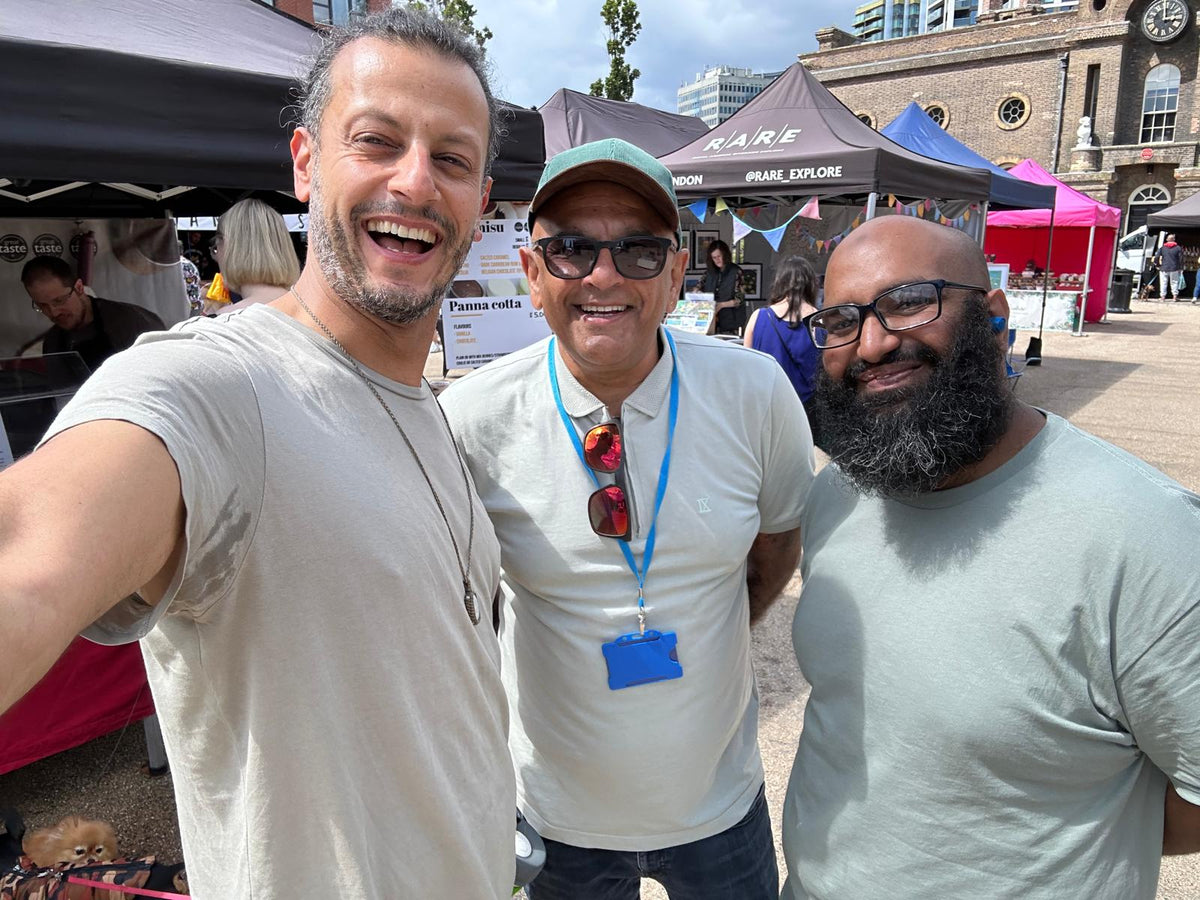Three men smiling and taking a selfie outdoors at what appears to be a market or event, with tents and a building in the background.