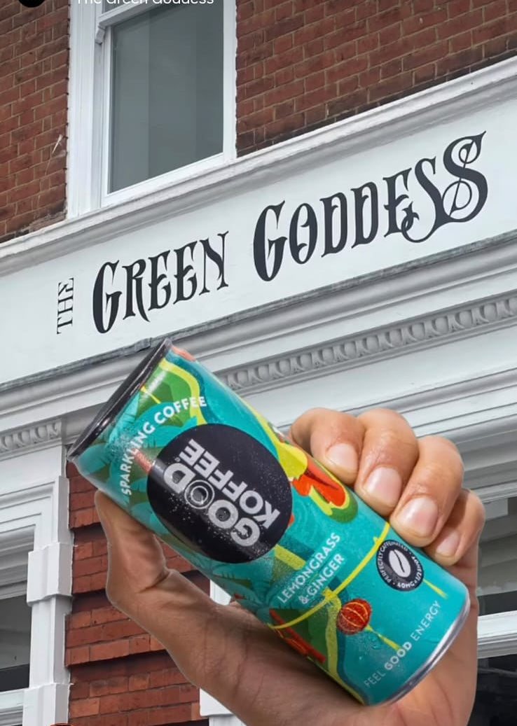 A hand holding a can of Good Coffee Roasted in front of a brick building with a white sign that reads 'The Green Goddess'.