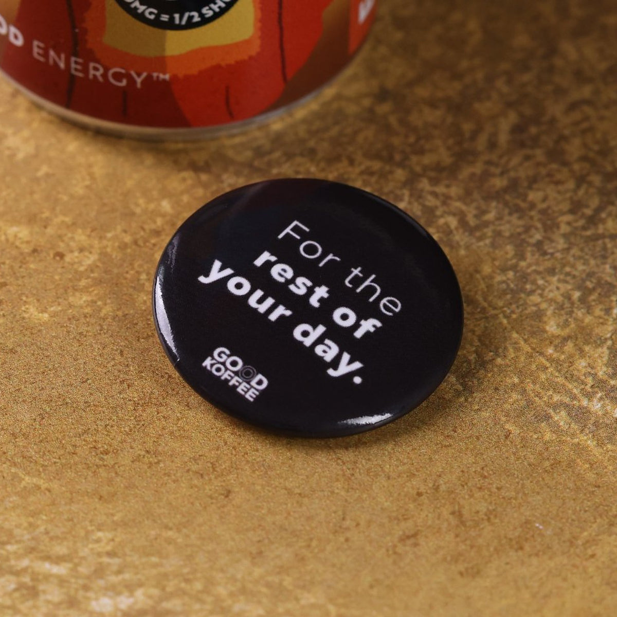 A round black coffee-themed pinback badge with the white text 'For the rest of your day.' and 'GOOD KOFFEE' logo, resting on a textured brown surface with a coffee can partially visible in the background.